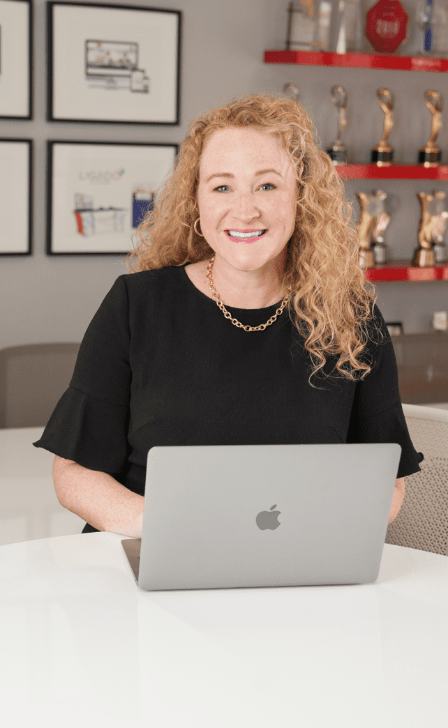Woman with curly blonde hair, wearing a black shirt and gold jewelry, using a laptop and smiling
