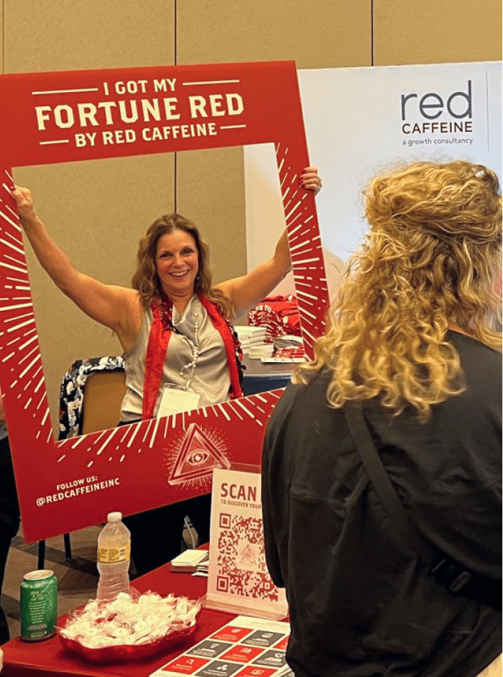 Woman in a conference setting, holding a big red picture frame that says "I Got My Fortune Red by Red Caffeine"