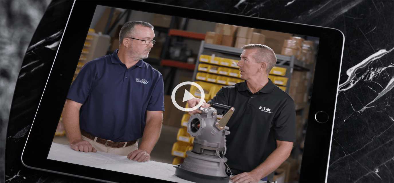 Tablet showing a still from a video, with 2 men in a warehouse talking about a mechanical part on the table in front of them