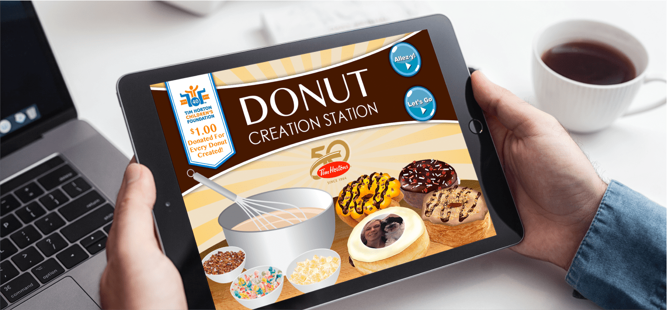 Hands holding a tablet next to a laptop and a coffee cup, with the tablet showing a "Donut Creation Station" app