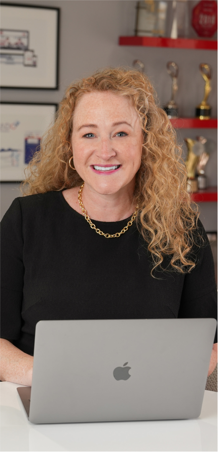 Woman with curly blonde hair, wearing a black shirt and gold jewelry, using a laptop and smiling
