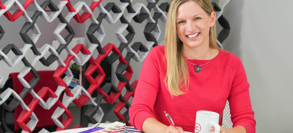 Woman with long blonde hair in a red shirt, smiling, at a table covered in colorful paint swatches and design papers