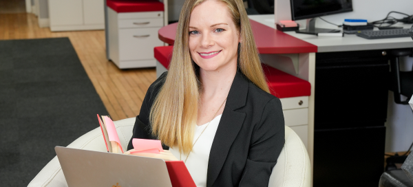 Woman with long blonde hair, wearing a black blazer, smiling as she uses a laptop and reads from a book with a red cover