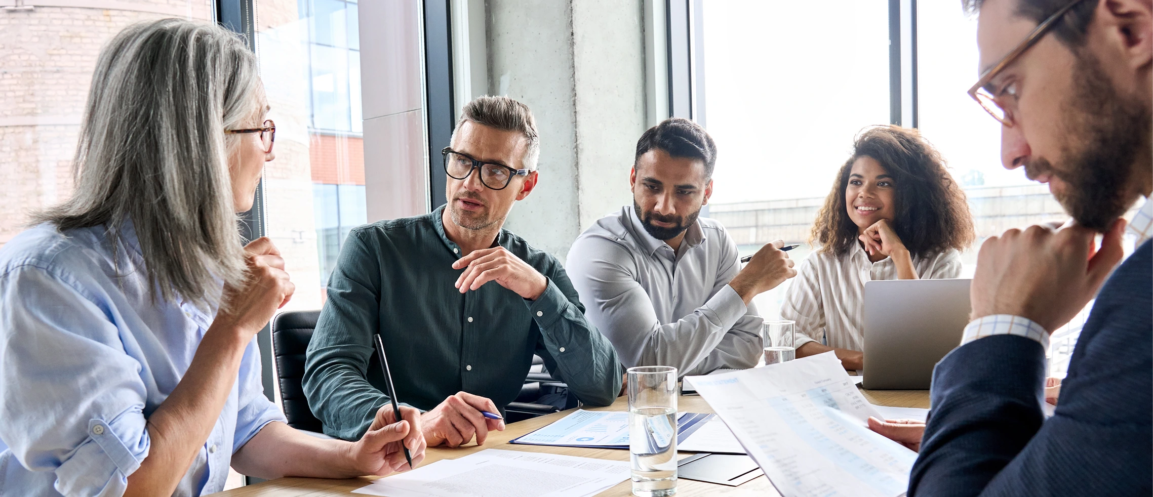 diverse-business-team-brainstorming Two men standing in a warehouse discuss information on a clipboard; one wears a business suit, the other wears a work jacket. Shelving and equipment are visible in the background.